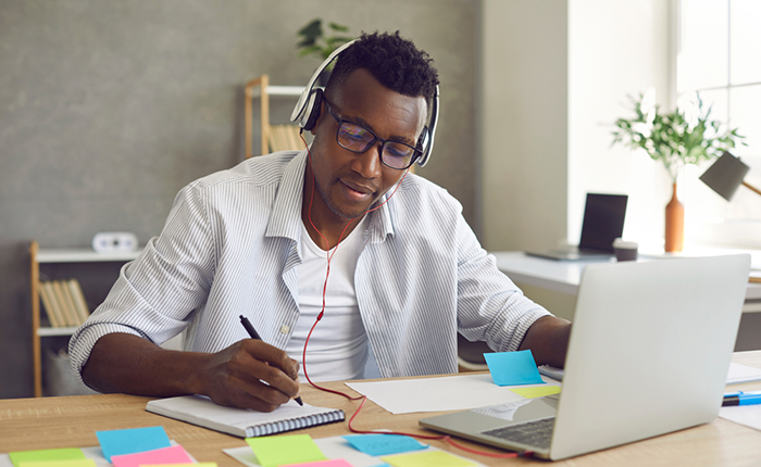 Man taking notes and wearing headphones while working on laptop