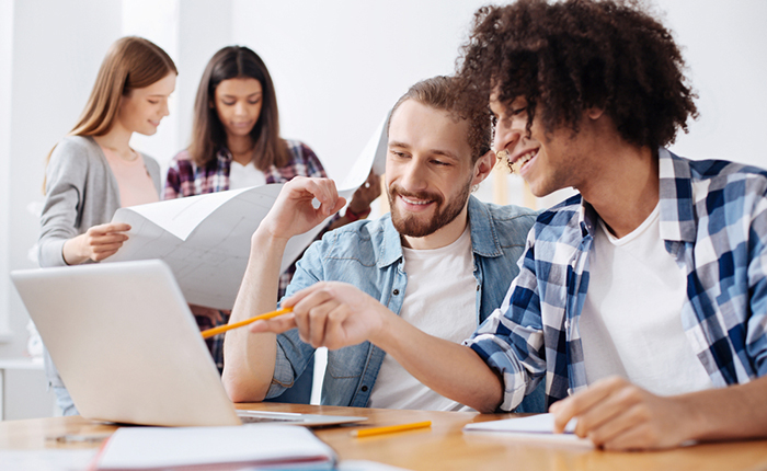 Two smiling men looking at a laptop with two women in the background looking at a large project plan schematic.