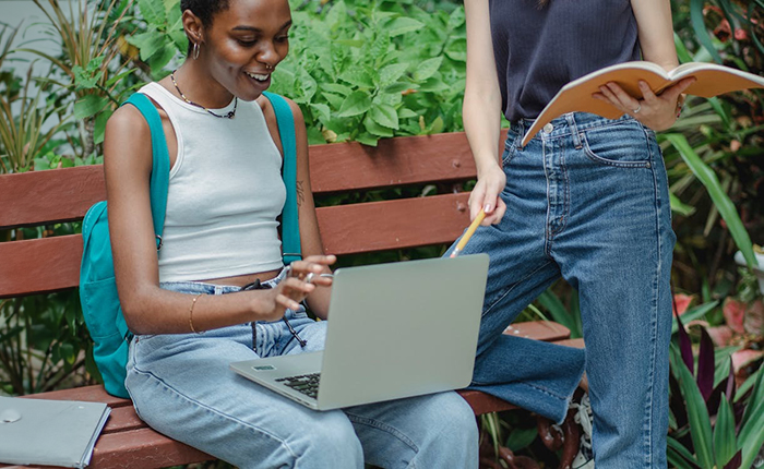 Smiling woman on a bench using a laptop with a friend pointing at the screen