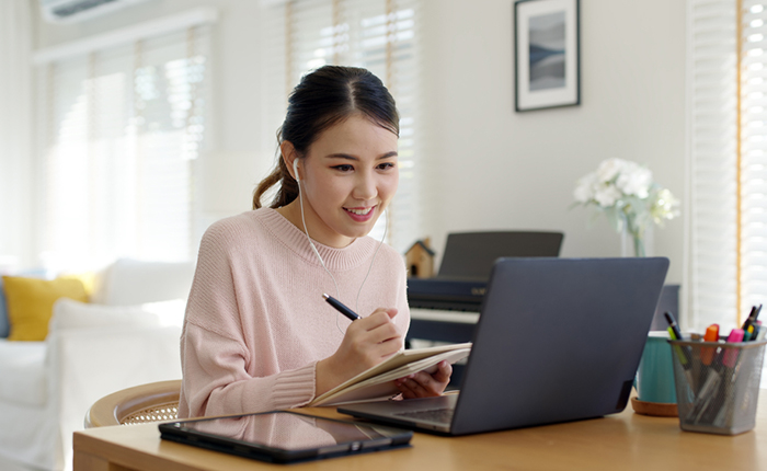 Woman wearing headphones listen to online course on her computer and taking notes.